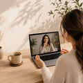 A woman sits at a wooden desk, seen from behind, interacting with a laptop screen during a video call. The scene is warmly lit with natural shadows from nearby plants, with a ceramic mug on the desk beside her.